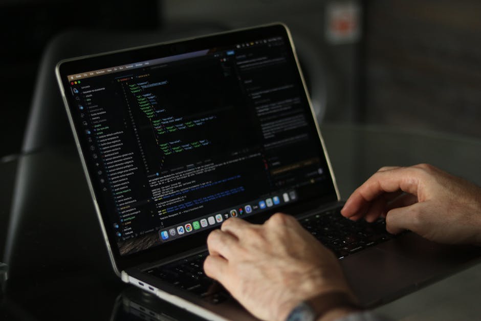 A close-up of a person’s hands typing on a sleek, silver laptop with a dark screen displaying lines of colorful code, positioned on a black table. In the background, there is a blurred dark interior